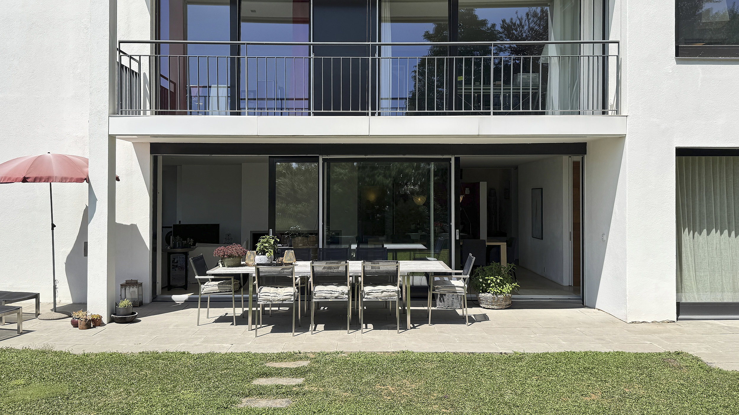 Covered garden terrace with dining area and lawn, white modern façade with balcony above